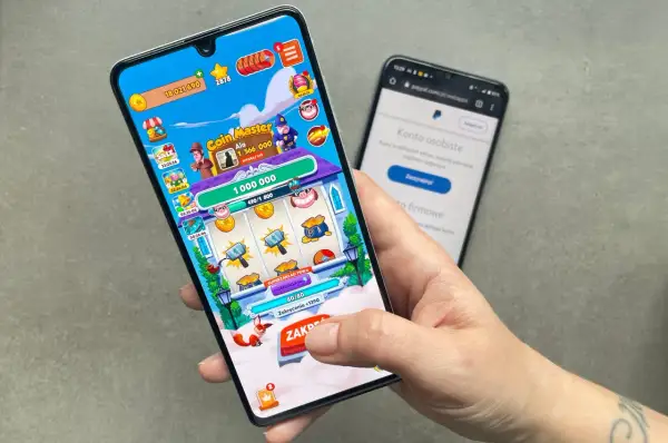 Dłoń trzymająca smartfon z grą Coin Master, obok drugi telefon z PayPal.