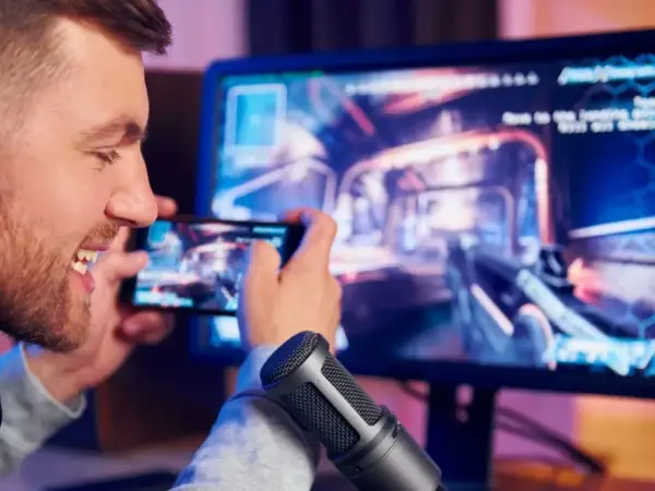 Mężczyzna gra w grę na telefonie, która jest wyświetlana na monitorze komputera.