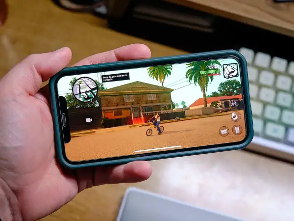 Ręka trzyma smartfon z grą RPG na telefon. Na ekranie widać postać na rowerze przed domem, palmy i ikony sterowania.
